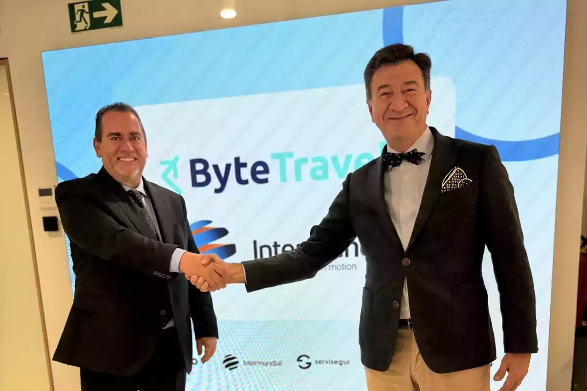 Dos hombres de negocios sonríen y se dan la mano frente a una pantalla con el logo de ByteTravel Dos hombres de negocios sonríen y se dan la mano frente a una pantalla con el logo de ByteTravel