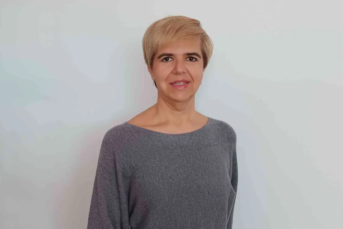 Mujer de cabello corto rubio con suéter gris posando frente a una pared blanca