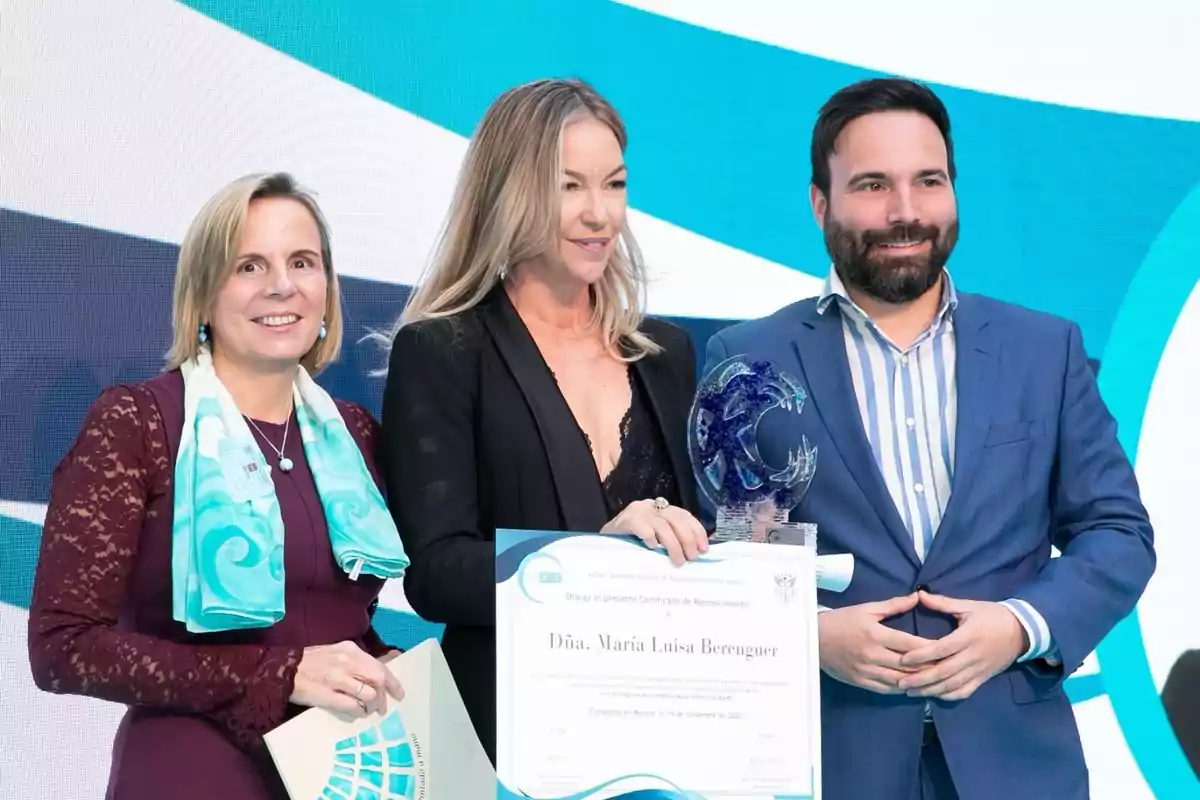 Tres personas posan sonrientes en un evento de premiación, una de ellas sostiene un diploma y un trofeo de cristal azul, mientras que las otras dos la acompañan en el escenario. Tres personas posan sonrientes en un evento de premiación, una de ellas sostiene un diploma y un trofeo de cristal azul, mientras que las otras dos la acompañan en el escenario.