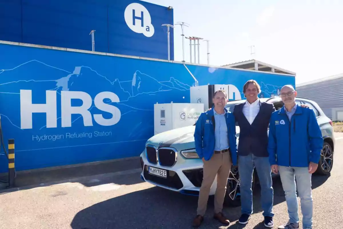 Tres hombres posan sonrientes frente a una estación de recarga de hidrógeno junto a un automóvil, con un mural azul de fondo que muestra las siglas HRS y equipo relacionado con hidrógeno