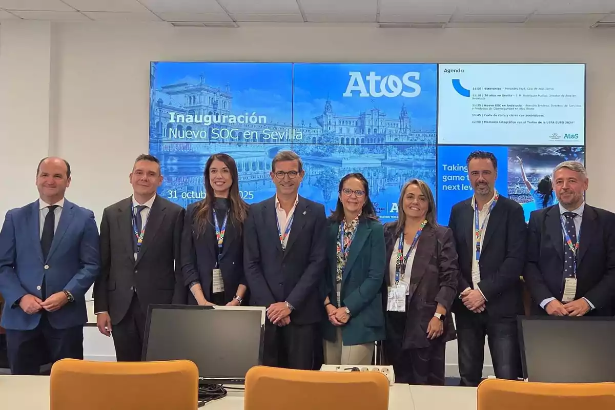 Ocho personas posan sonrientes en una sala de oficina frente a una pantalla grande que muestra el logo de Atos y el anuncio de la inauguración de un nuevo SOC en Sevilla Ocho personas posan sonrientes en una sala de oficina frente a una pantalla grande que muestra el logo de Atos y el anuncio de la inauguración de un nuevo SOC en Sevilla