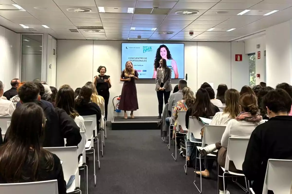 Audiencia sentada en una sala de conferencias mientras tres mujeres están de pie en el escenario frente a una pantalla grande que muestra información del evento