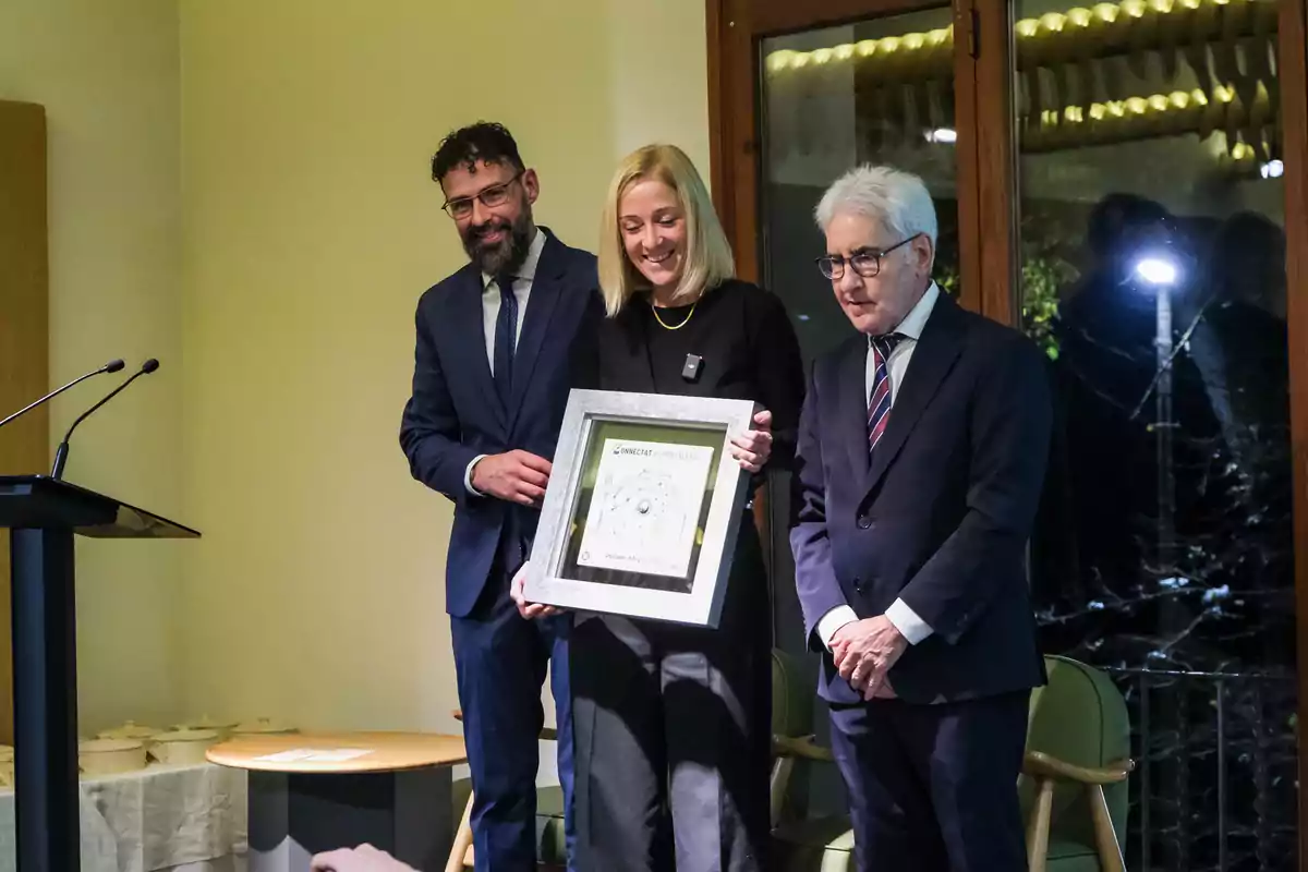 Tres personas vestidas de manera formal posan sonrientes mientras una de ellas sostiene un cuadro con un reconocimiento en el centro de un salón iluminado Tres personas vestidas de manera formal posan sonrientes mientras una de ellas sostiene un cuadro con un reconocimiento en el centro de un salón iluminado