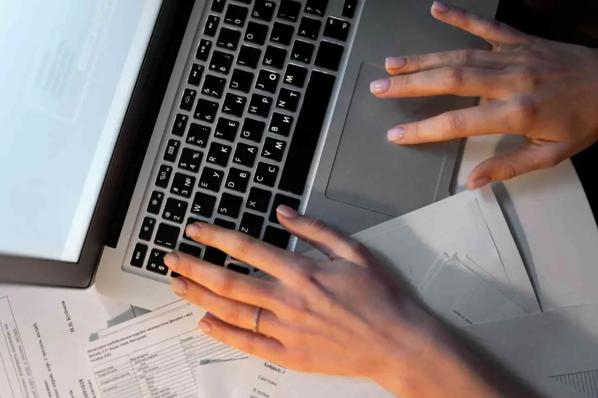 Manos escribiendo en una computadora portátil sobre una mesa con documentos y papeles.