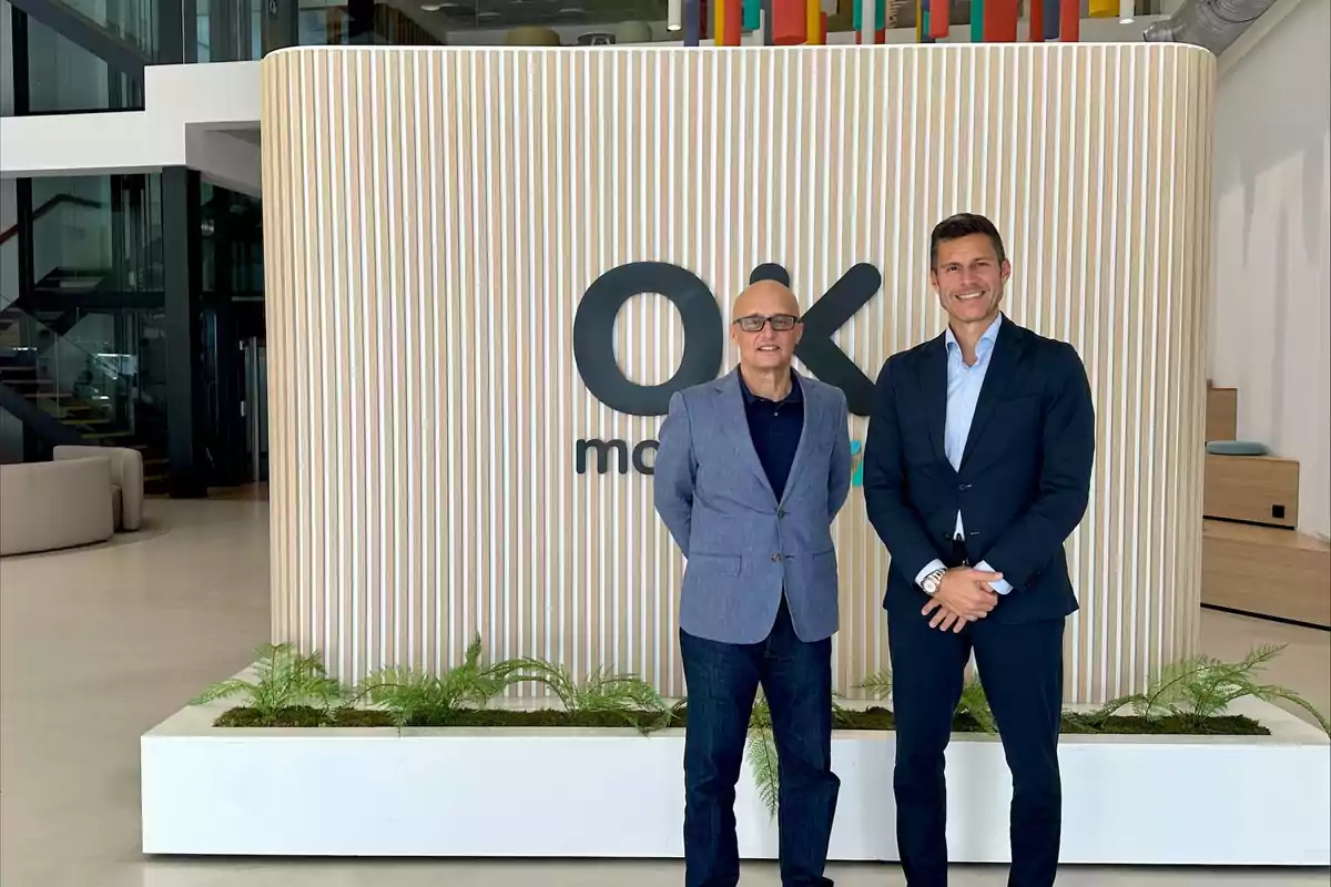 Dos hombres de negocios posan sonrientes frente a un letrero de OK Mobility en una oficina moderna y luminosa
