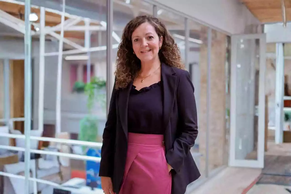 Mujer de cabello rizado y castaño, vestida con saco negro y falda fucsia, sonríe mientras posa en un entorno de oficina moderno con paredes de vidrio y plantas al fondo