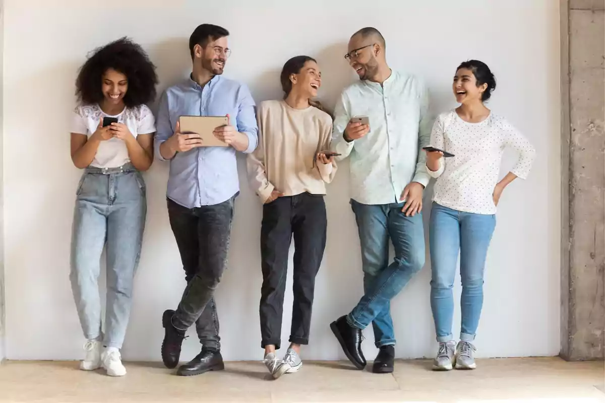 Cinco personas jóvenes de pie junto a una pared blanca, sonriendo y conversando mientras usan dispositivos electrónicos.