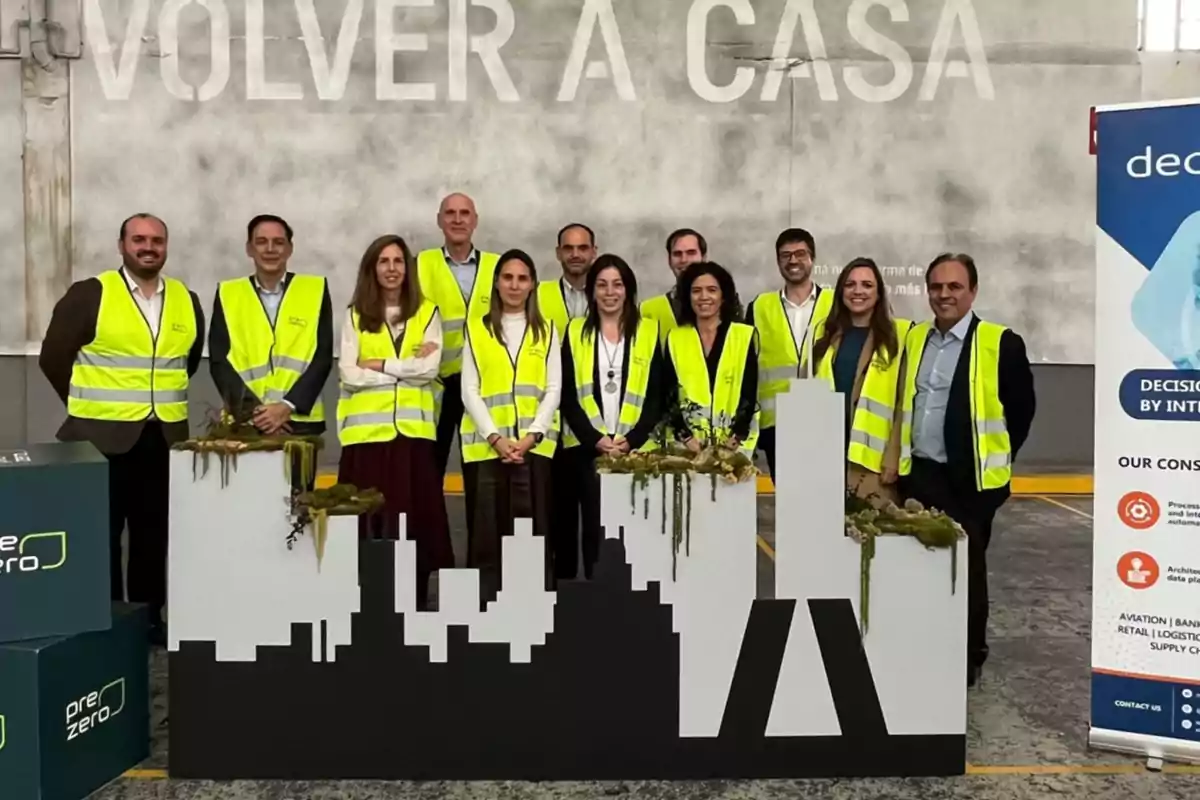 Grupo de personas con chalecos reflectantes posando en un entorno industrial junto a una figura de ciudad y un cartel promocional de servicios de consultoría Grupo de personas con chalecos reflectantes posando en un entorno industrial junto a una figura de ciudad y un cartel promocional de servicios de consultoría