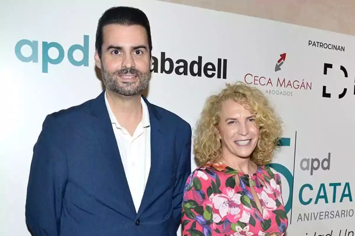 Un hombre con traje azul y una mujer con vestido floral posan sonrientes frente a un fondo con logotipos de empresas.