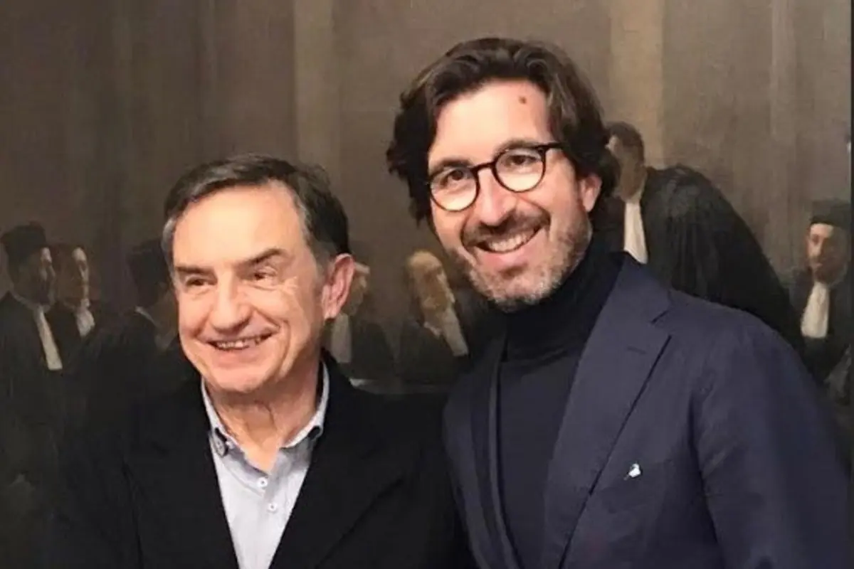 Dos hombres sonrientes posan juntos frente a una pintura de fondo donde se observan varias personas vestidas con togas negras