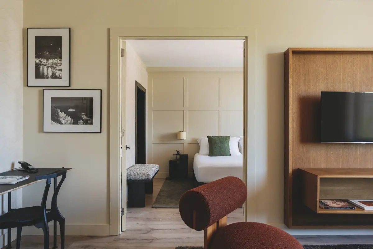 Habitación de hotel moderna con sala de estar y dormitorio conectados, decoración minimalista en tonos neutros, cama con cojín verde, escritorio con teléfono, televisor de pantalla plana en mueble de madera y fotografías en blanco y negro en la pared