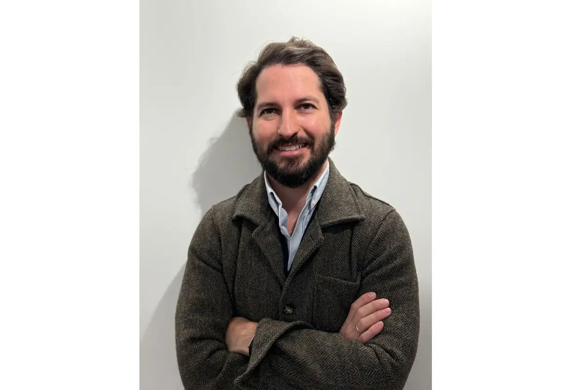 Hombre de cabello y barba oscuros con chaqueta marrón y camisa clara posando con los brazos cruzados frente a una pared blanca Hombre de cabello y barba oscuros con chaqueta marrón y camisa clara posando con los brazos cruzados frente a una pared blanca