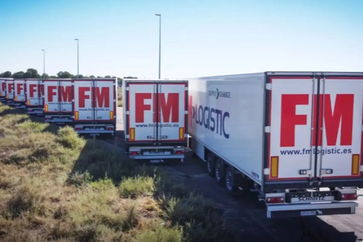Varios camiones de la empresa FM Logistic estacionados en fila junto a un camino rodeado de vegetación.