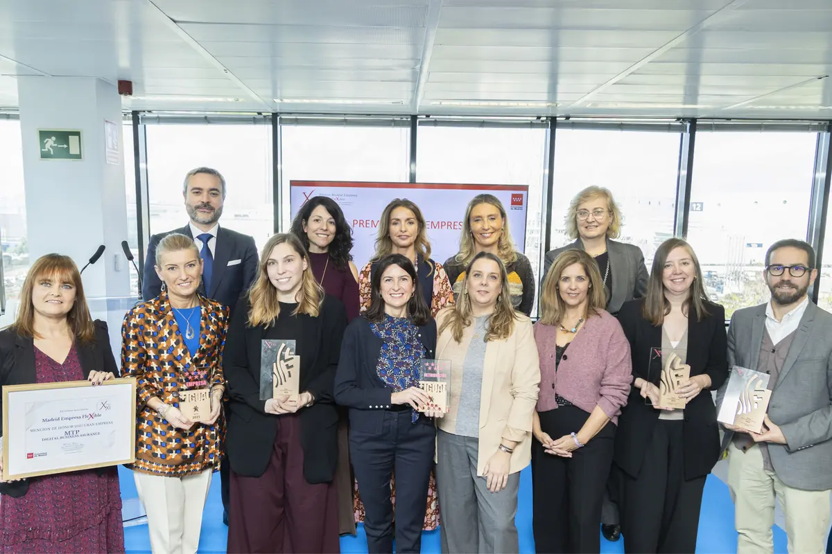 Grupo de personas posando en un evento de premiación empresarial, varias de ellas sostienen trofeos y un diploma, todos sonríen y están en un entorno de oficina moderno con ventanales al fondo.