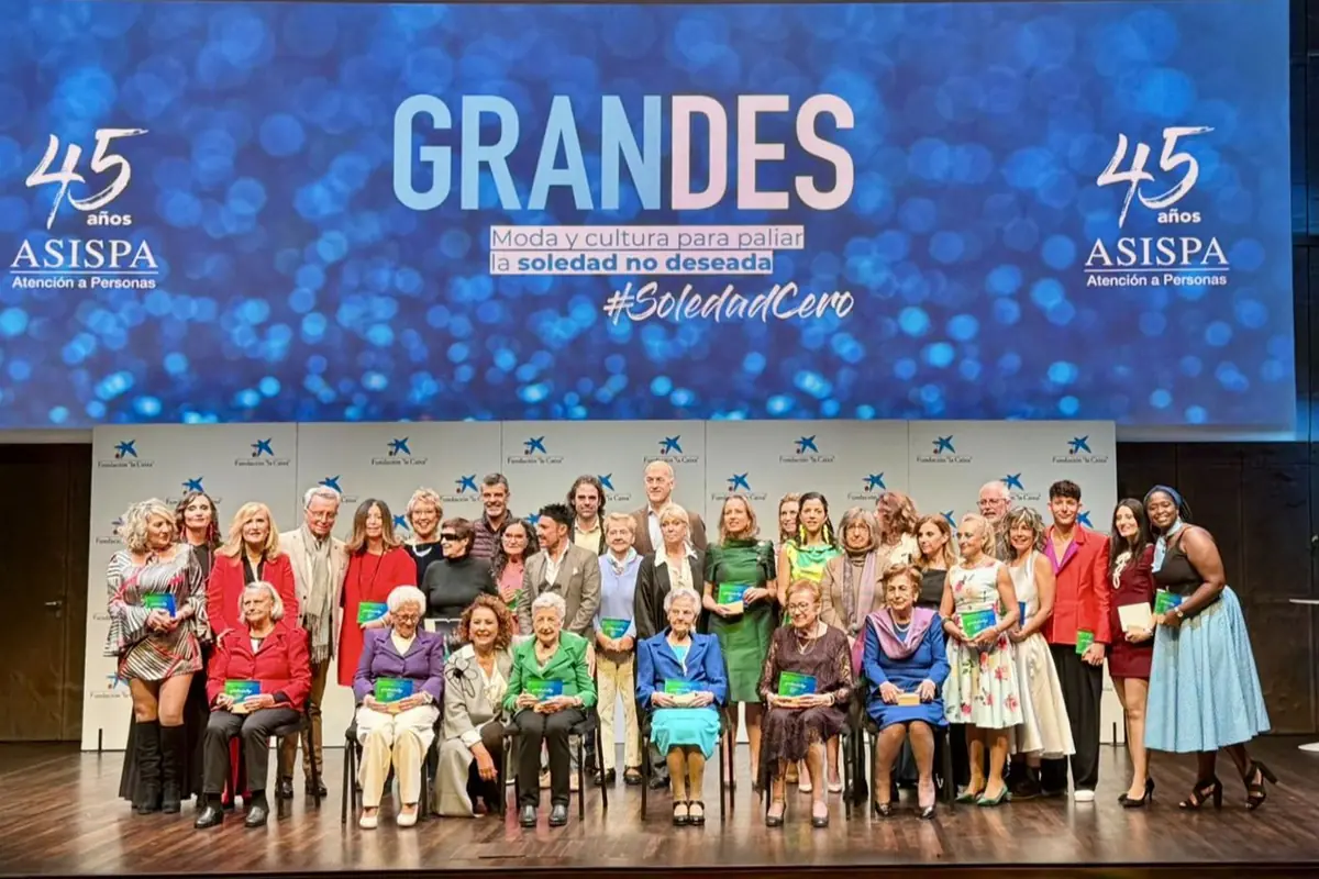 Grupo de personas de diferentes edades posando en un escenario durante un evento con un gran cartel azul de fondo que dice GRANDES y promueve la cultura y la moda para combatir la soledad no deseada Grupo de personas de diferentes edades posando en un escenario durante un evento con un gran cartel azul de fondo que dice GRANDES y promueve la cultura y la moda para combatir la soledad no deseada