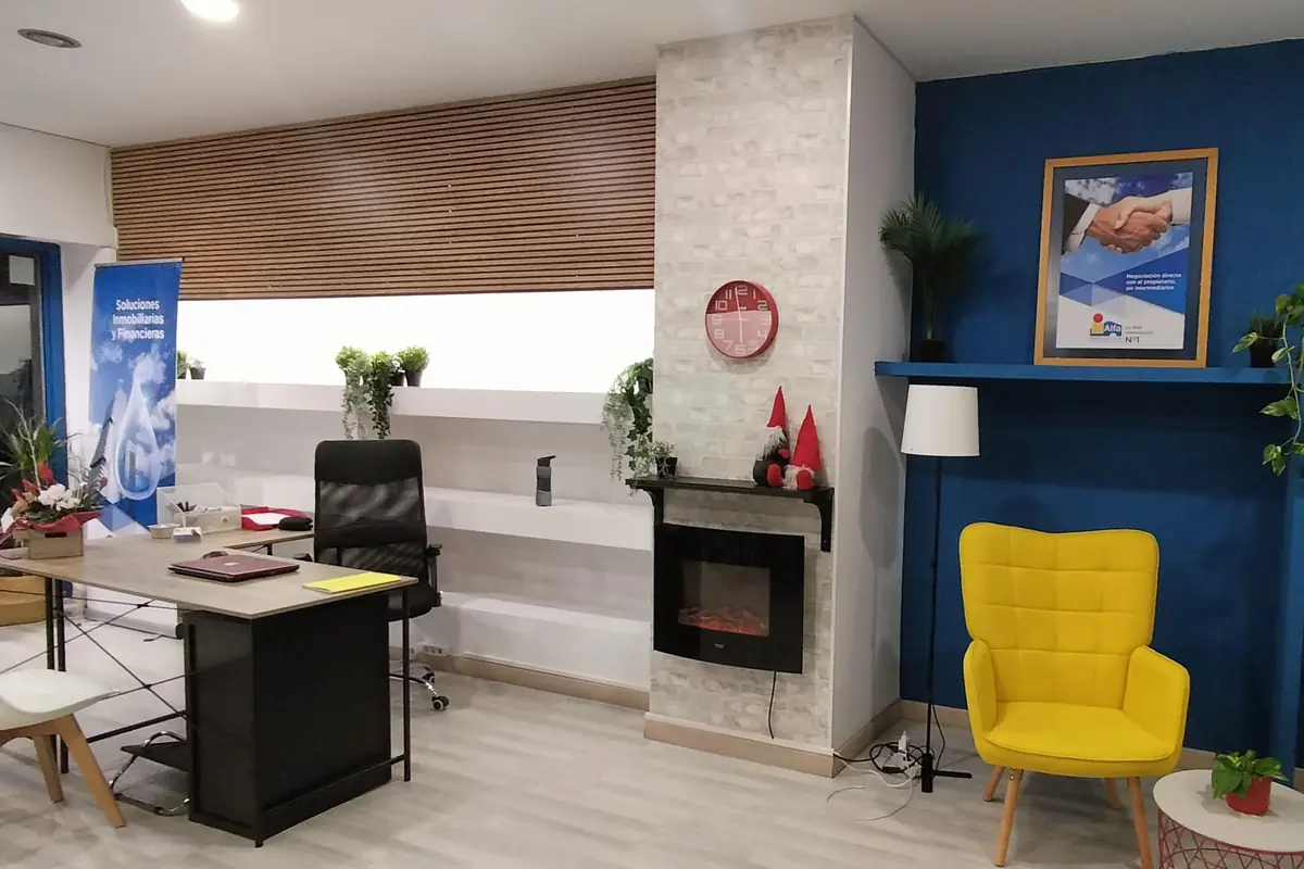Oficina moderna con escritorio negro, silla ejecutiva, estanterías blancas con plantas, chimenea decorativa, pared azul con cuadro corporativo y sillón amarillo junto a una lámpara de pie