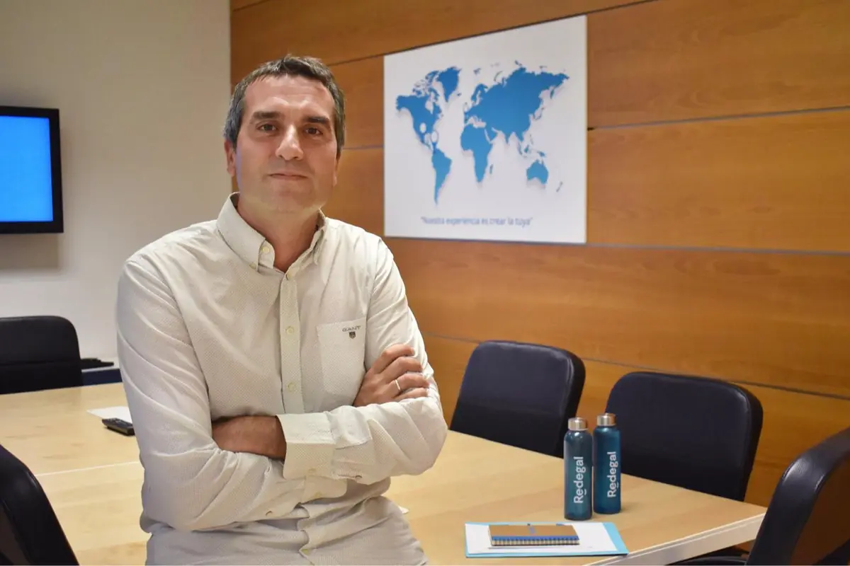 Hombre sentado en una sala de reuniones con una mesa, dos botellas azules, un cuaderno y un mapa del mundo en la pared de fondo