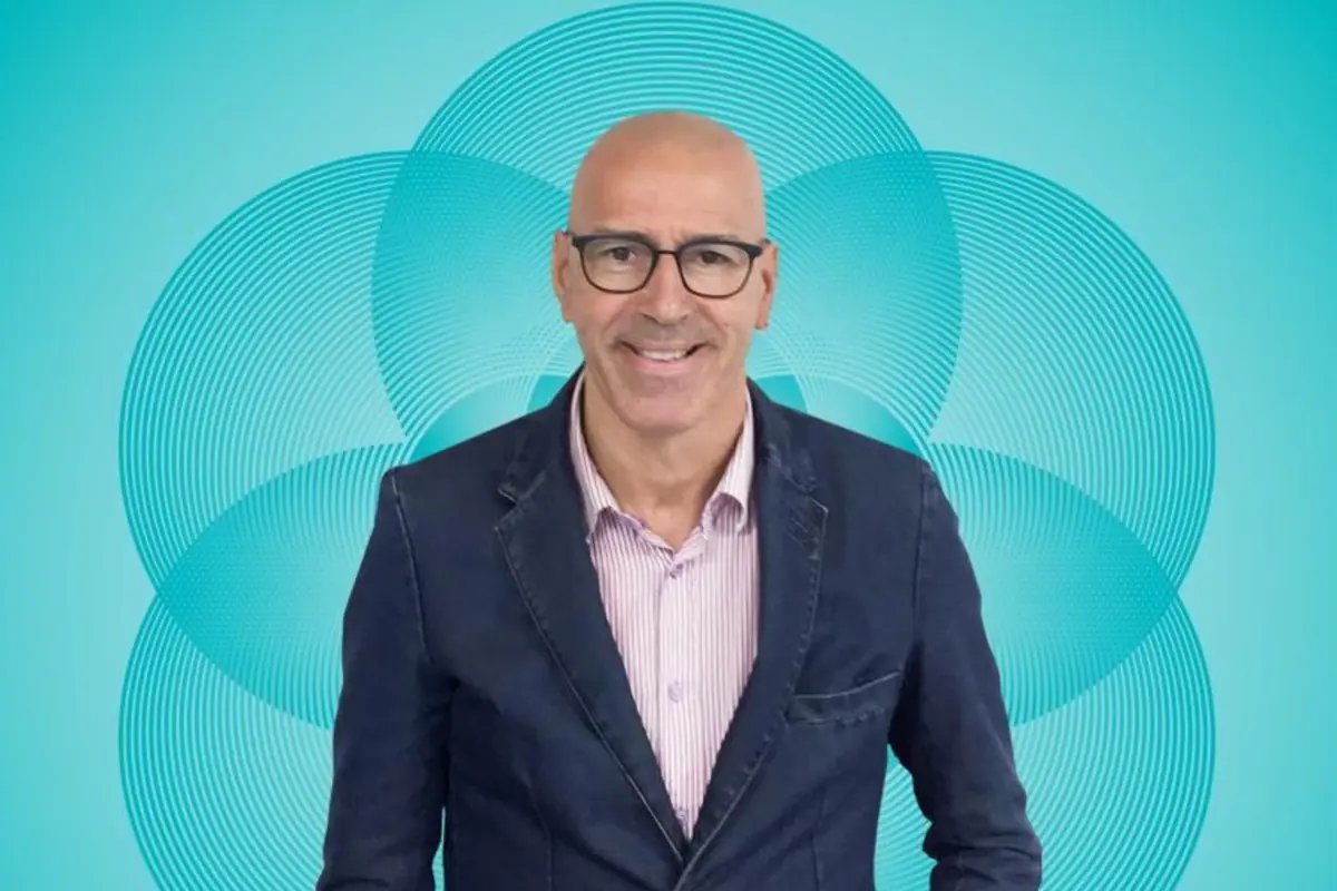 Hombre calvo con gafas y chaqueta azul sonriendo frente a un fondo turquesa con círculos concéntricos