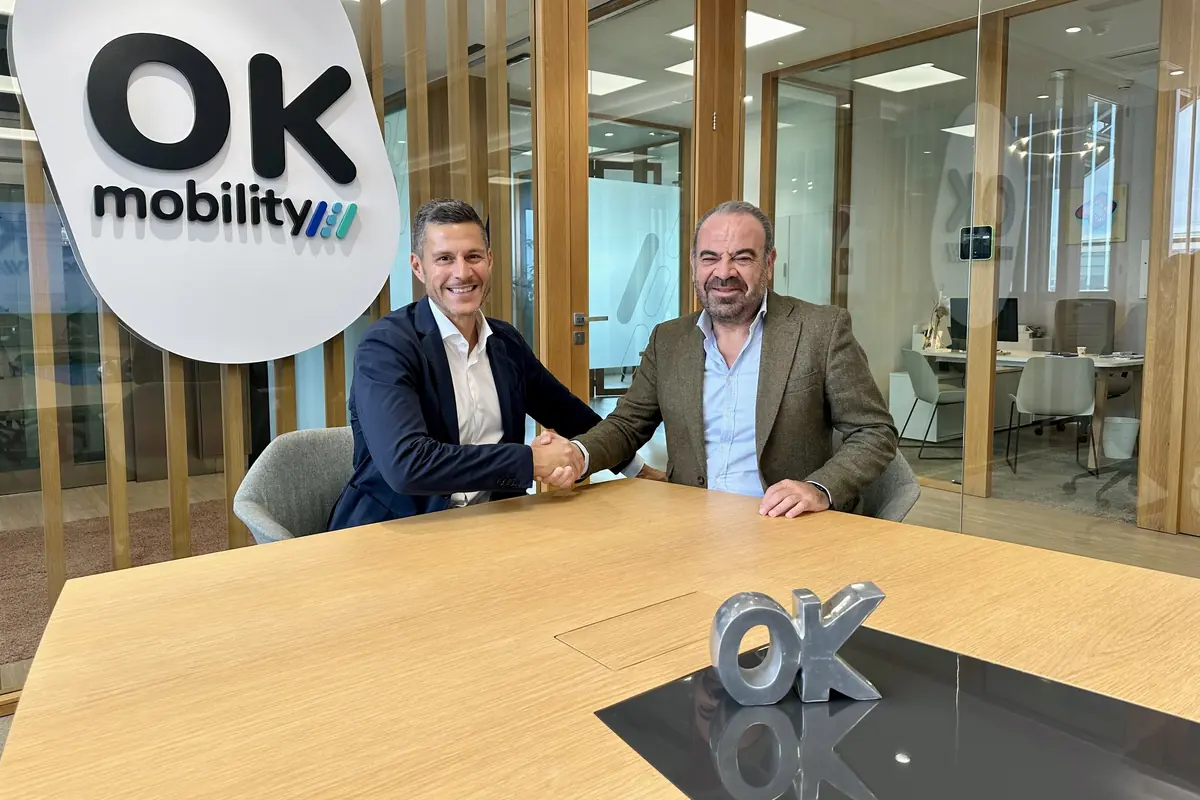 Dos hombres de negocios se dan la mano en una sala de reuniones moderna de OK Mobility con el logotipo de la empresa visible en la pared y sobre la mesa