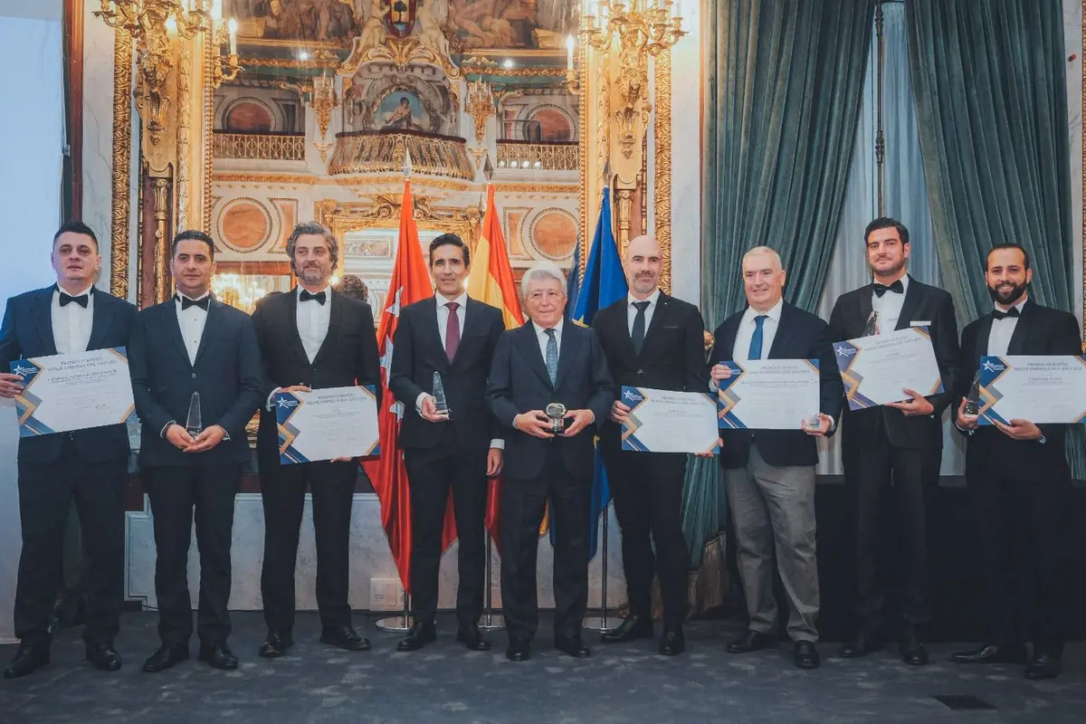 Ocho hombres vestidos de traje posan en fila sosteniendo diplomas y trofeos en una elegante sala decorada con cortinas verdes, espejos y lámparas de araña, con banderas oficiales al fondo. Ocho hombres vestidos de traje posan en fila sosteniendo diplomas y trofeos en una elegante sala decorada con cortinas verdes, espejos y lámparas de araña, con banderas oficiales al fondo.