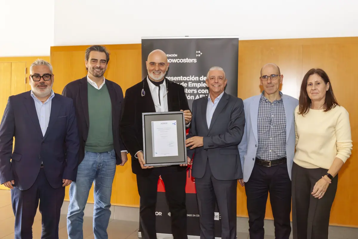 Seis personas posan sonrientes en un acto institucional mientras el hombre del centro sostiene un diploma enmarcado frente a un cartel corporativo de fondo
