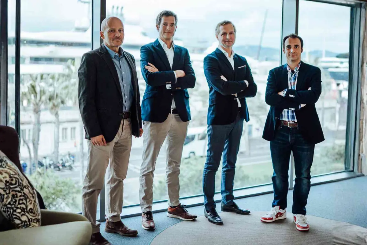 Cuatro hombres de negocios posan de pie frente a grandes ventanales con vista urbana, vestidos con americana y pantalones informales