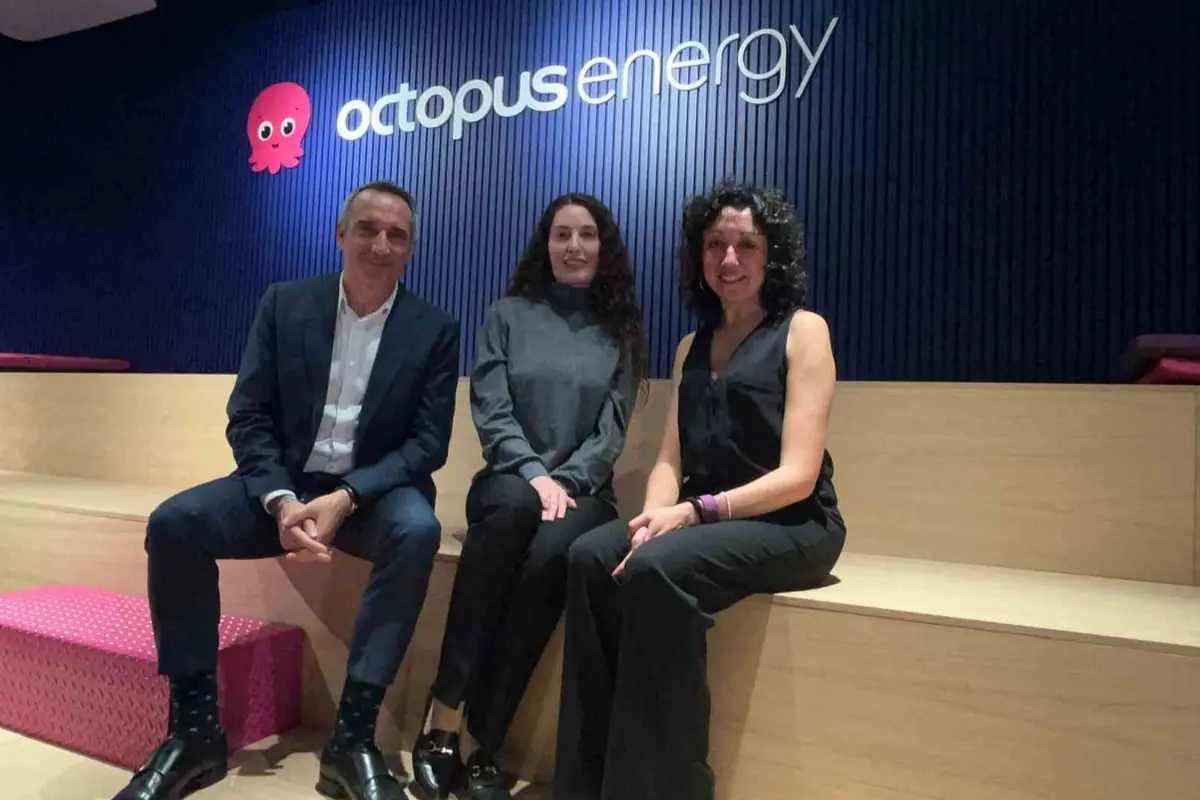 Tres personas sentadas en gradas de madera en una oficina con el logo de Octopus Energy en la pared azul detrás de ellas Tres personas sentadas en gradas de madera en una oficina con el logo de Octopus Energy en la pared azul detrás de ellas