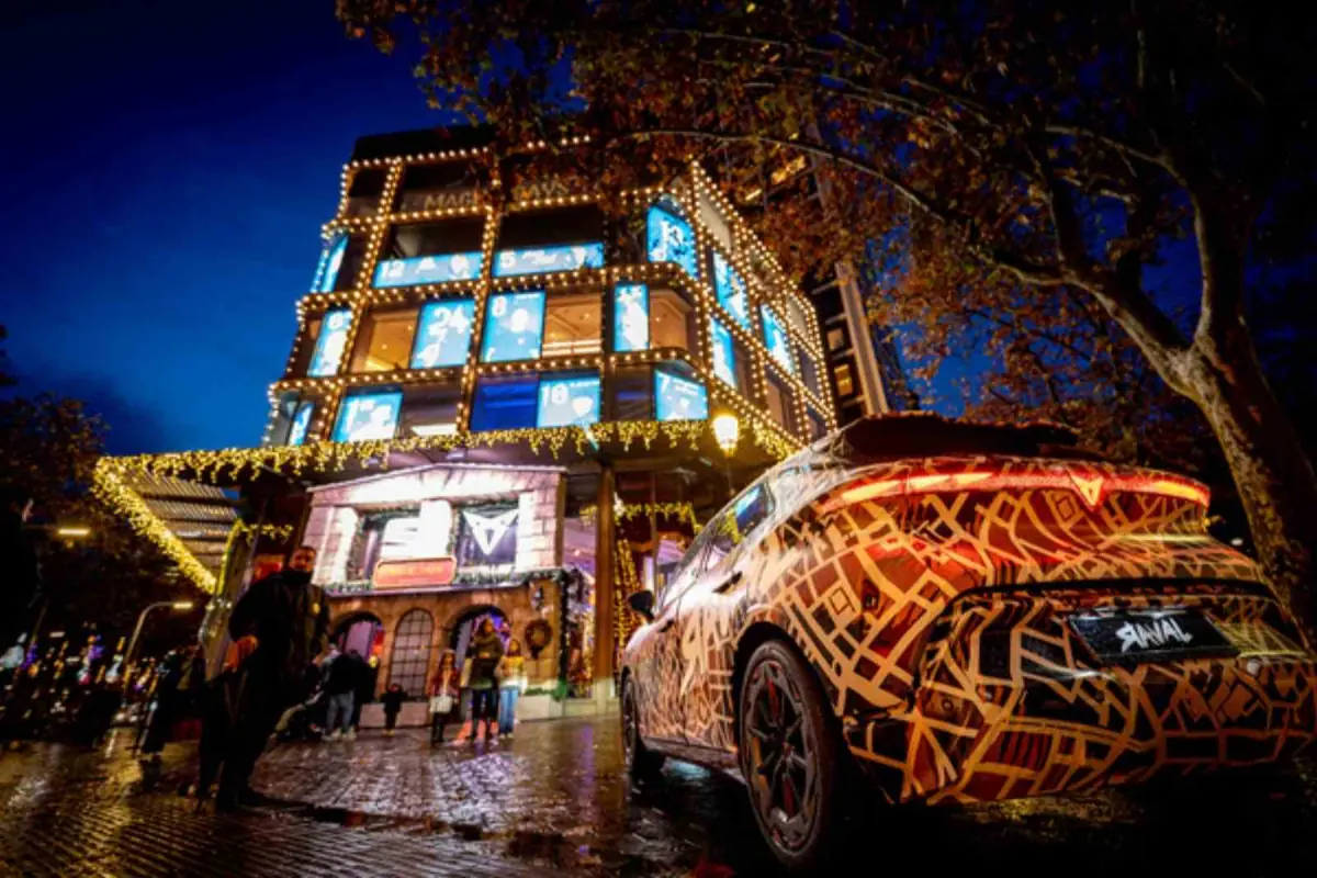 Coche con vinilo de diseño urbano aparcado frente a un edificio iluminado con luces y pantallas navideñas en una calle mojada al anochecer
