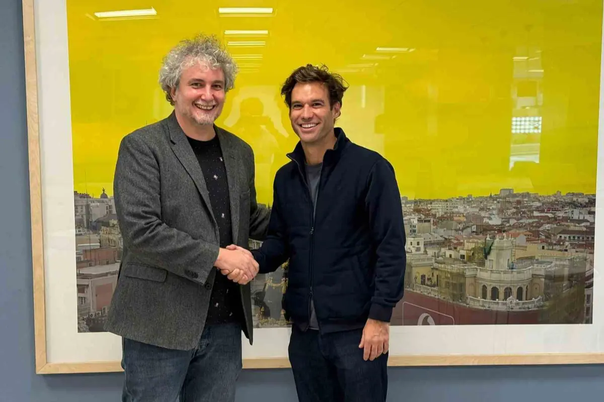 Dos hombres sonrientes se dan la mano frente a una gran fotografía de una ciudad con fondo amarillo brillante