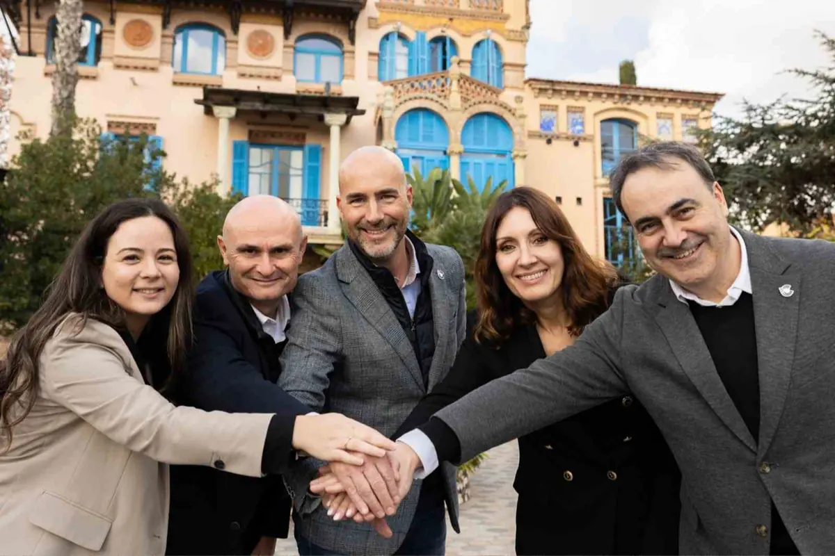 Cinco personas sonrientes con las manos unidas en gesto de trabajo en equipo frente a un edificio histórico con ventanas azules