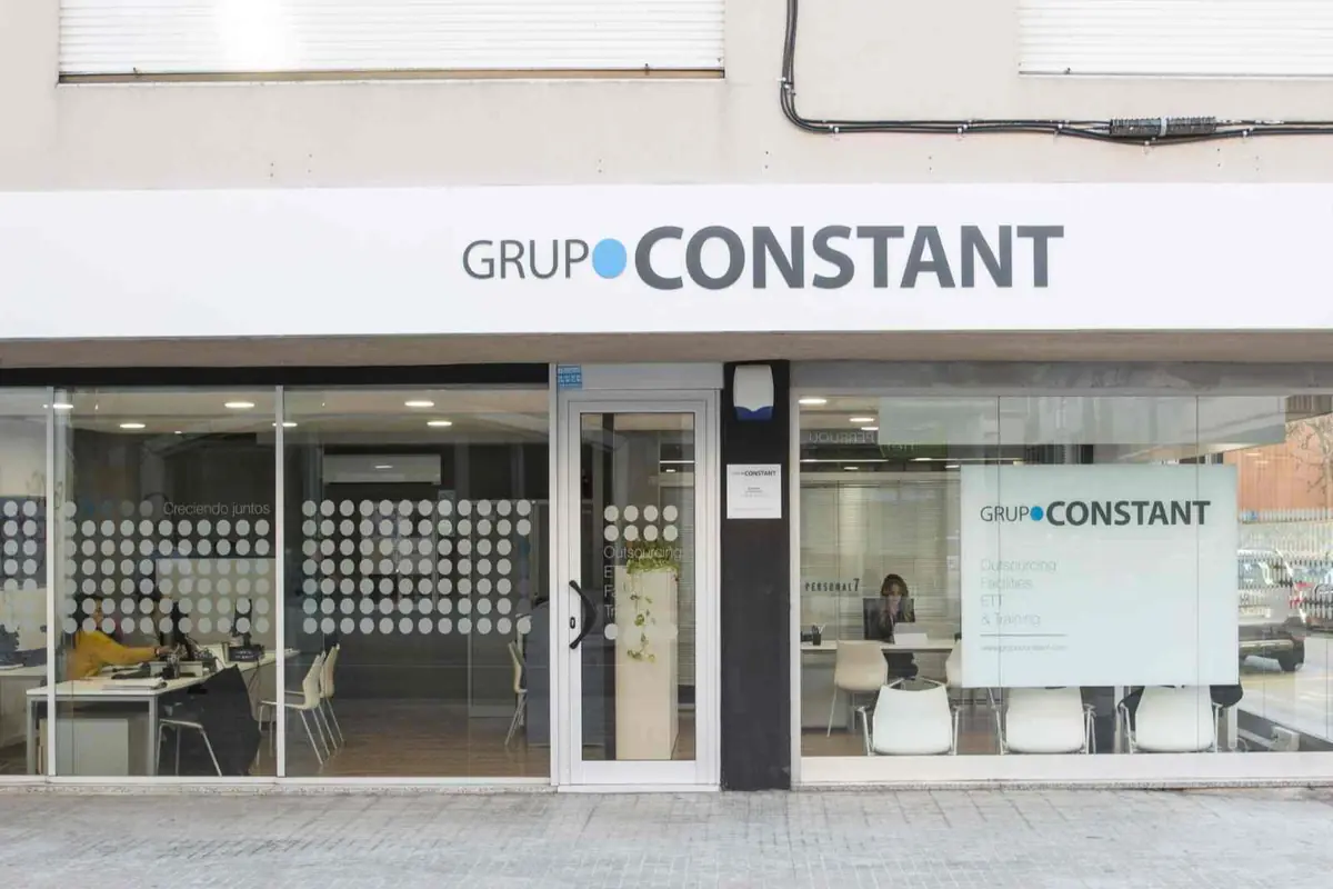 Fachada acristalada de una oficina de Grupo Constant con rótulo corporativo y personas trabajando en el interior Fachada acristalada de una oficina de Grupo Constant con rótulo corporativo y personas trabajando en el interior