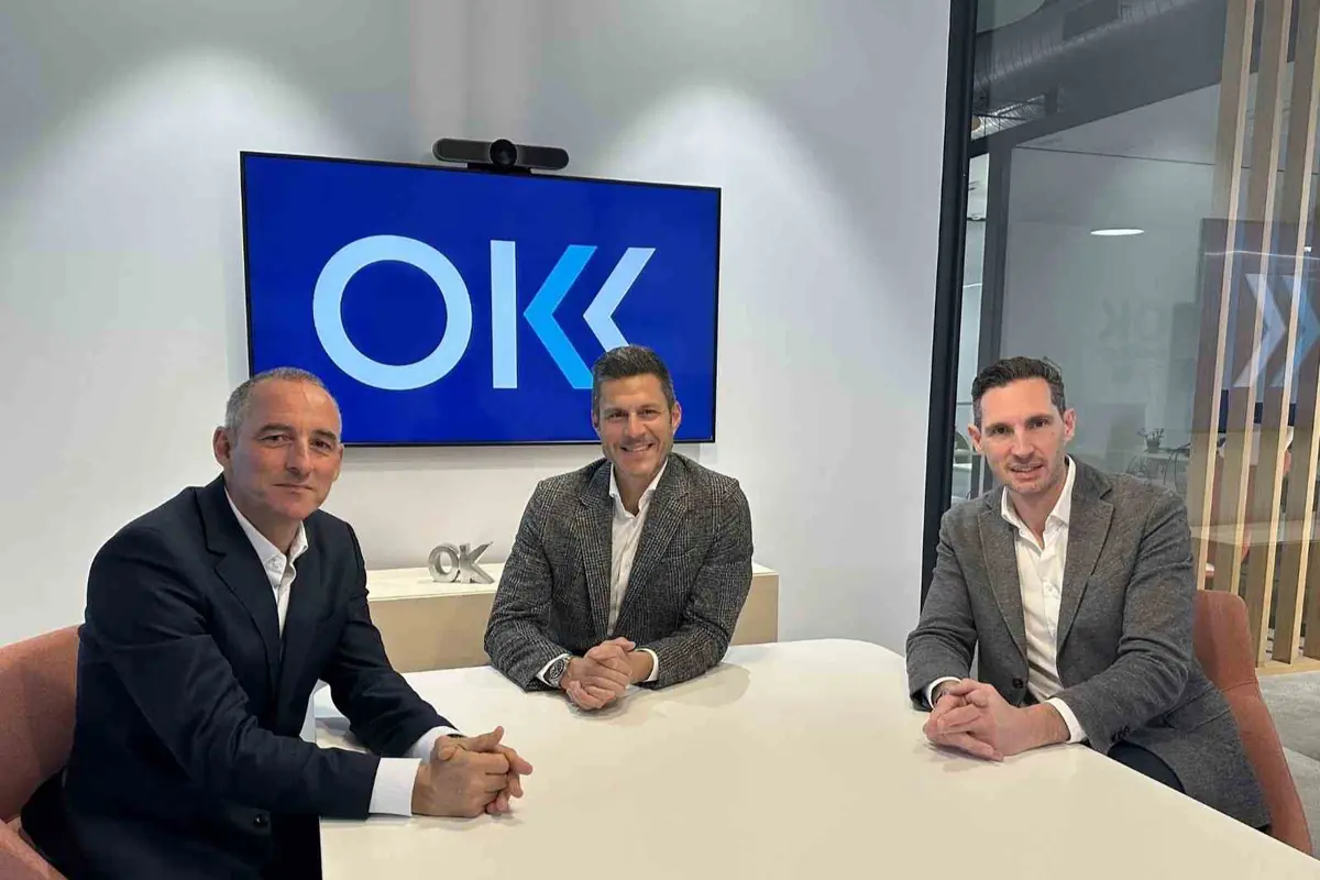 Tres hombres de negocios sentados en una sala de reuniones moderna frente a una pantalla con el logotipo azul de OK