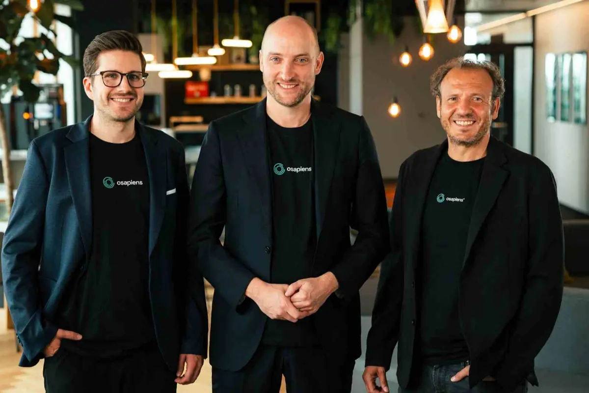 Tres hombres sonriendo con camisetas negras de la marca osapiens y chaquetas oscuras posando en una oficina moderna iluminada con lámparas colgantes