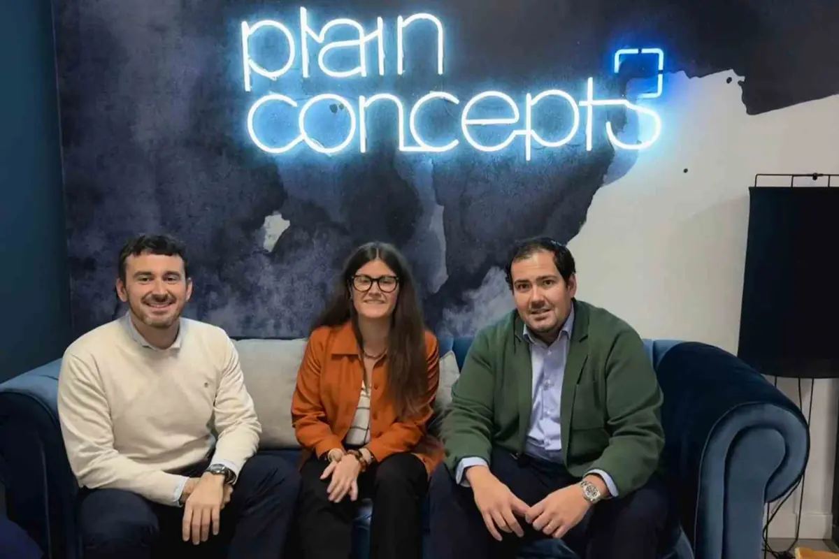 Tres personas sentadas en un sofá azul sonriendo frente a una pared oscura con el letrero de neón encendido que dice plain concepts Tres personas sentadas en un sofá azul sonriendo frente a una pared oscura con el letrero de neón encendido que dice plain concepts