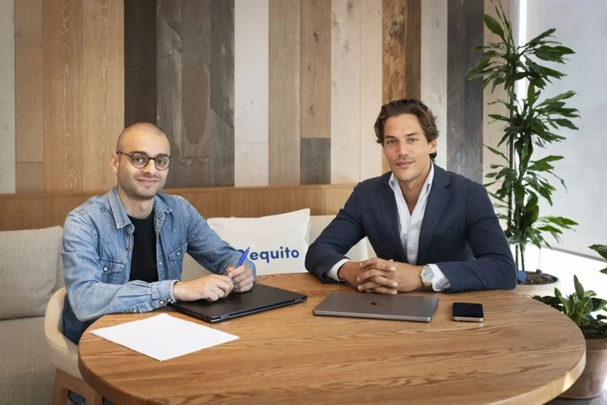 Dos hombres jóvenes sentados en una mesa redonda de madera en una oficina moderna con portátiles documentos y plantas al fondo