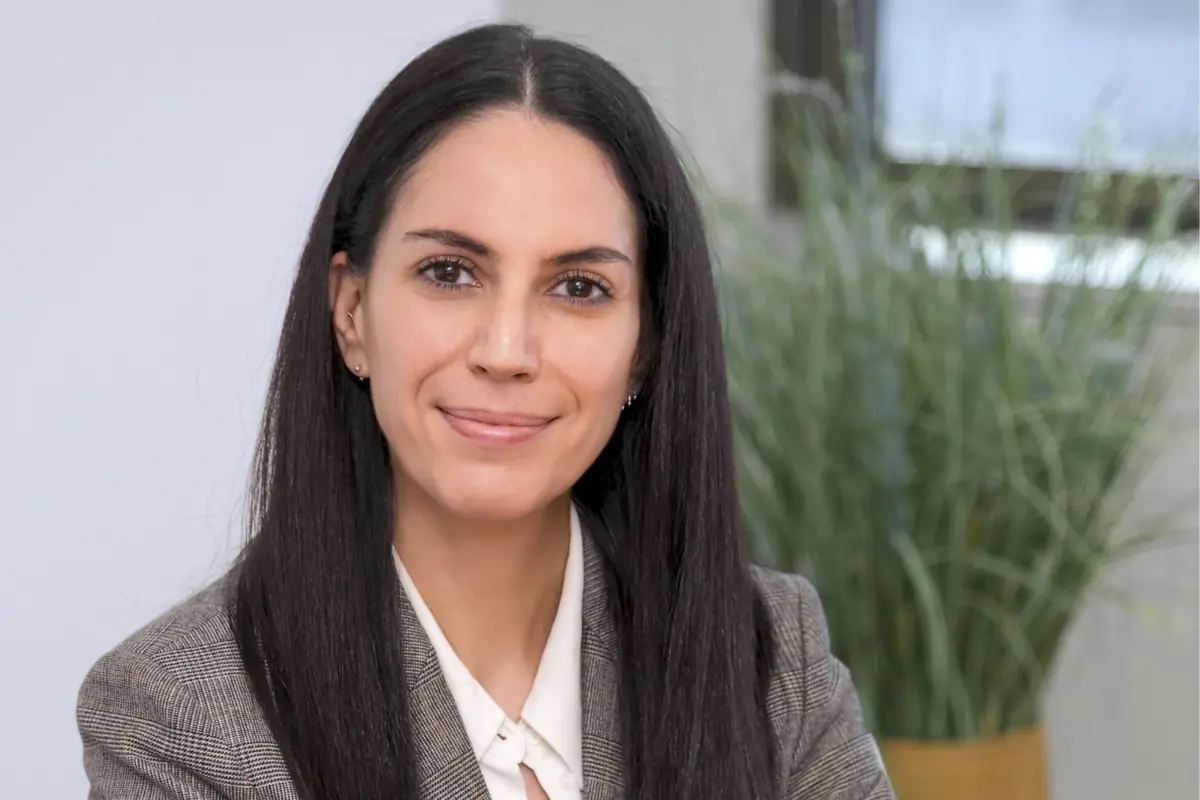 Retrato de una mujer joven con el pelo largo y liso sonriendo levemente y vistiendo americana gris y blusa blanca en una oficina con planta verde de fondo