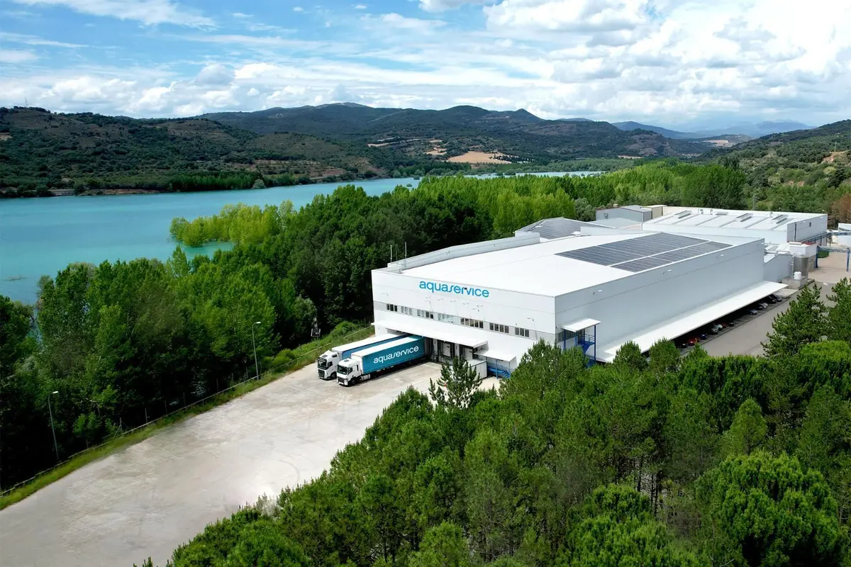 Vista aérea de una planta industrial de Aquaservice rodeada de árboles junto a un gran embalse de agua turquesa y montañas al fondo