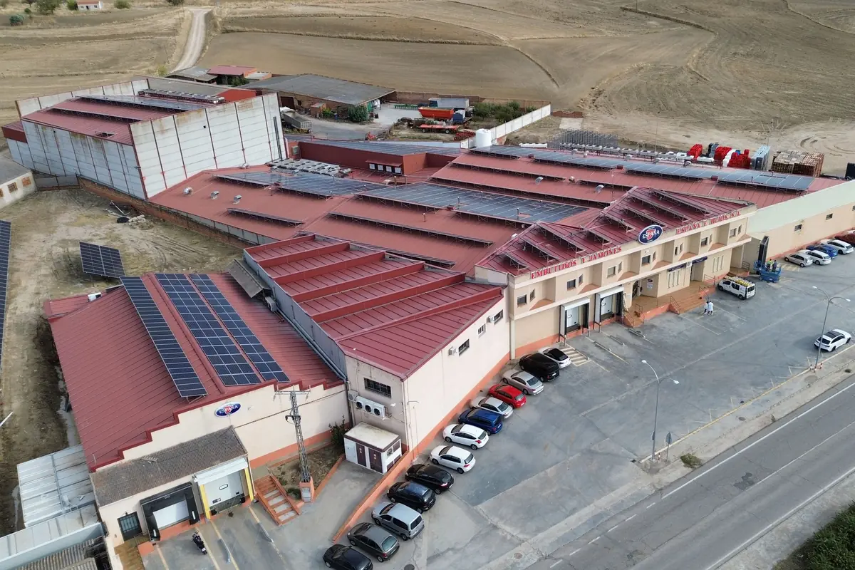 Vista aérea de una nave industrial con tejado rojo equipada con paneles solares y un aparcamiento con varios coches en una zona rural
