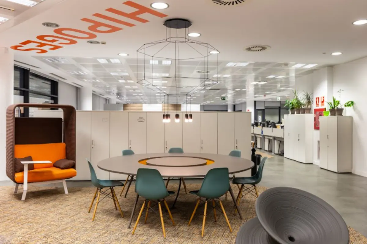 Oficina moderna y luminosa con mesa redonda de reuniones, sillas verdes, sillón naranja tipo cabina y armarios blancos al fondo Oficina moderna y luminosa con mesa redonda de reuniones, sillas verdes, sillón naranja tipo cabina y armarios blancos al fondo