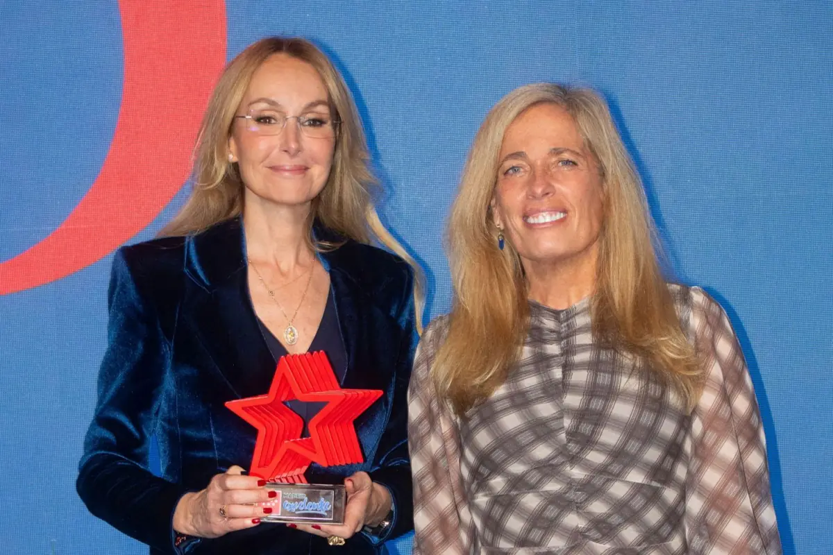 Dos mujeres rubias posan sonrientes frente a un fondo azul mientras una de ellas sostiene un trofeo rojo en forma de estrella