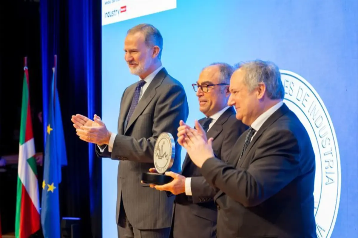 Tres hombres con traje en un escenario aplauden mientras uno de ellos sostiene un trofeo redondo frente a un fondo azul con banderas y un logotipo institucional