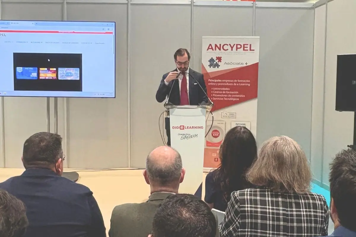 Hombre con traje y corbata roja dando una conferencia sobre e-learning en un atril frente a un público sentado, con una pantalla y un cartel de ANCYPEL al fondo