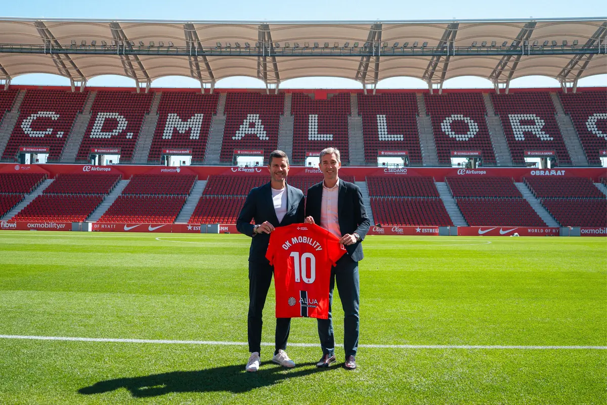 Dos hombres trajeados posan en el césped del estadio del RCD Mallorca sosteniendo una camiseta roja con el número 10 y el patrocinio OK Mobility frente a las gradas vacías
