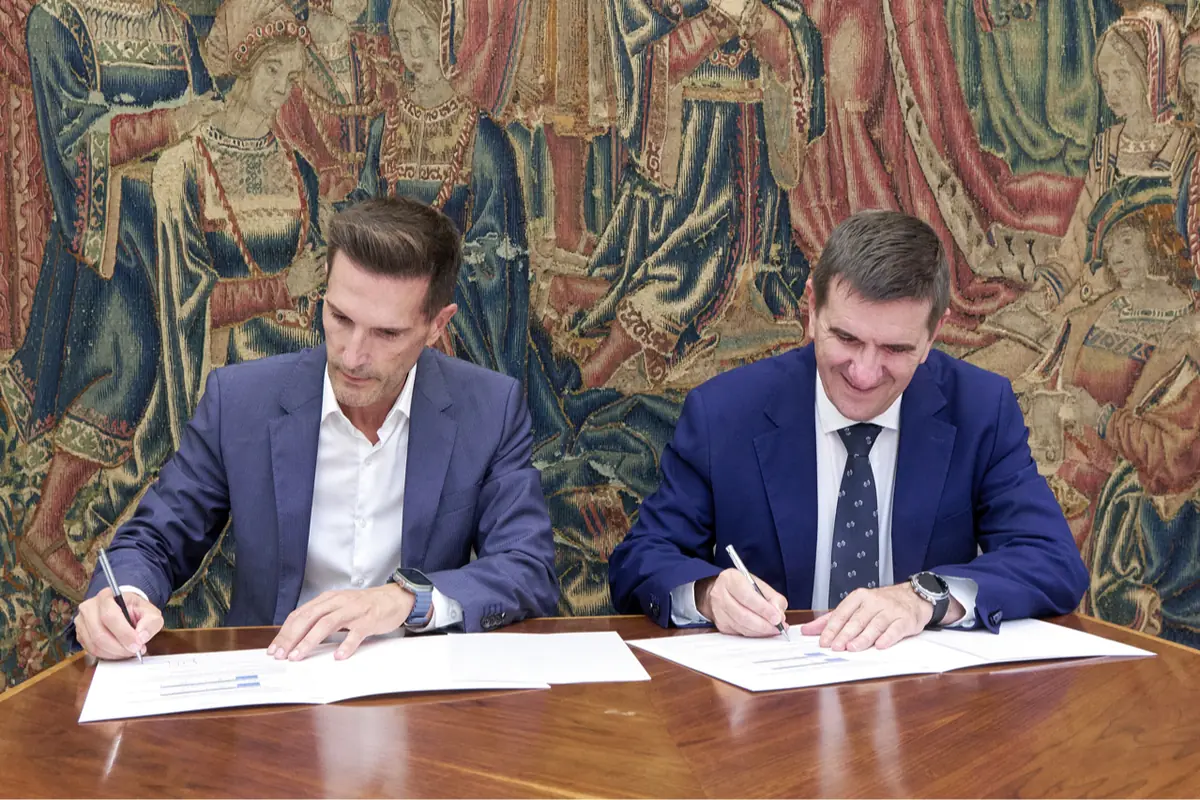 Dos hombres con traje firmando documentos sobre una mesa de madera frente a un tapiz antiguo con figuras históricas