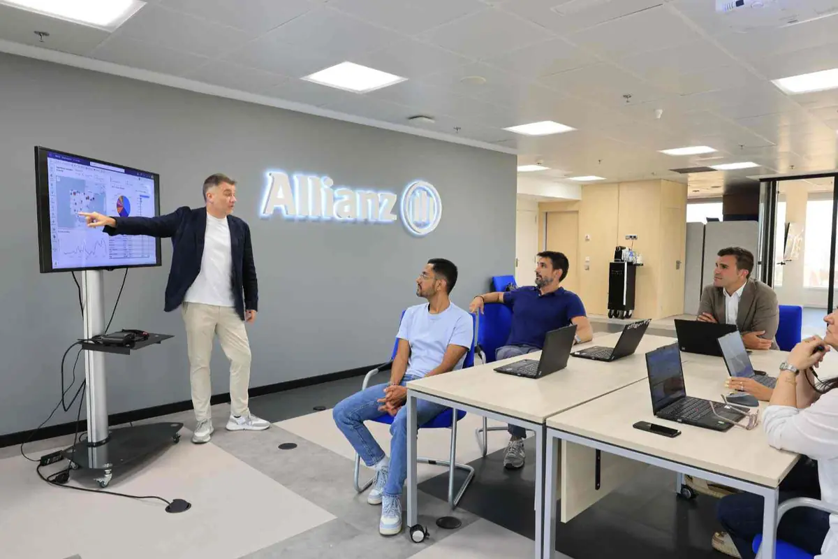 Hombre de pie señalando una pantalla con gráficos mientras cuatro personas con portátiles lo observan en una sala de reuniones de la empresa Allianz