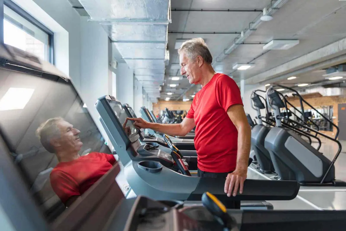 Hombre mayor con camiseta roja usando la pantalla de una cinta de correr en un gimnasio moderno