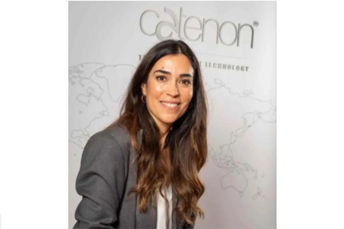 Mujer joven con chaqueta gris sonriendo frente a un fondo corporativo con mapa del mundo y el logotipo de una empresa Mujer joven con chaqueta gris sonriendo frente a un fondo corporativo con mapa del mundo y el logotipo de una empresa