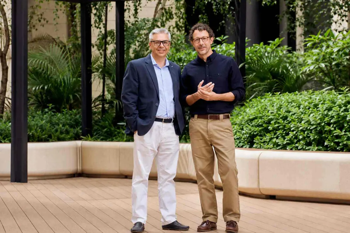 Dos hombres de pie en una terraza de madera rodeada de plantas y vegetación, vestidos de manera informal elegante y mirando a la cámara