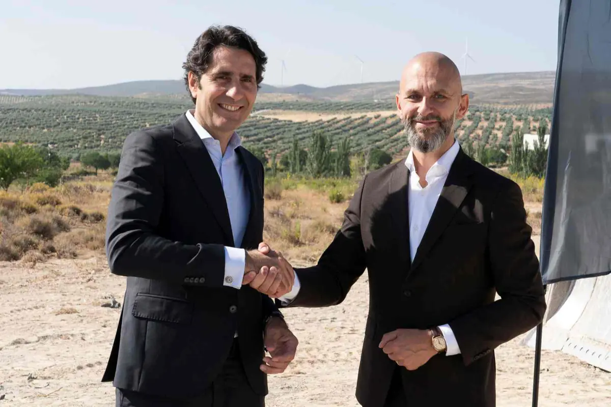 Dos hombres de negocios con traje oscuro se dan la mano al aire libre frente a un paisaje rural con colinas y aerogeneradores al fondo