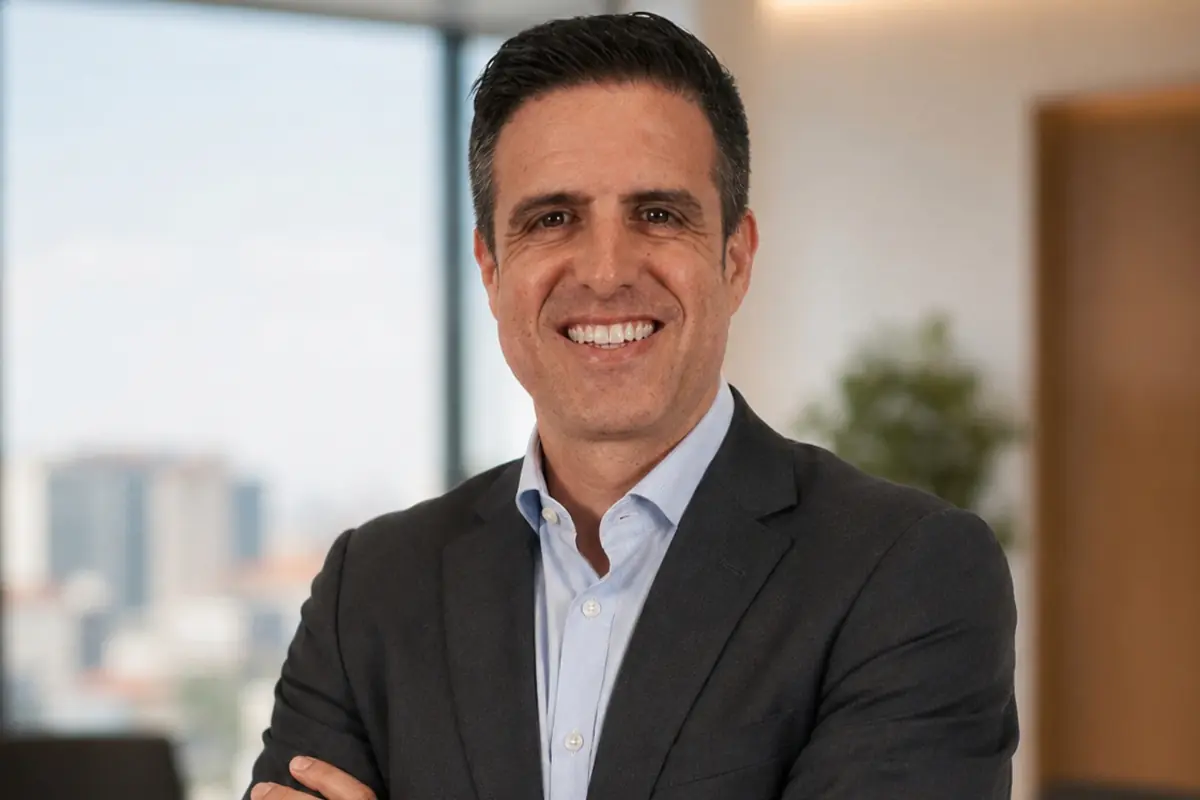 Hombre de mediana edad con traje oscuro y camisa clara sonriendo con los brazos cruzados en una oficina luminosa con ventanales al fondo